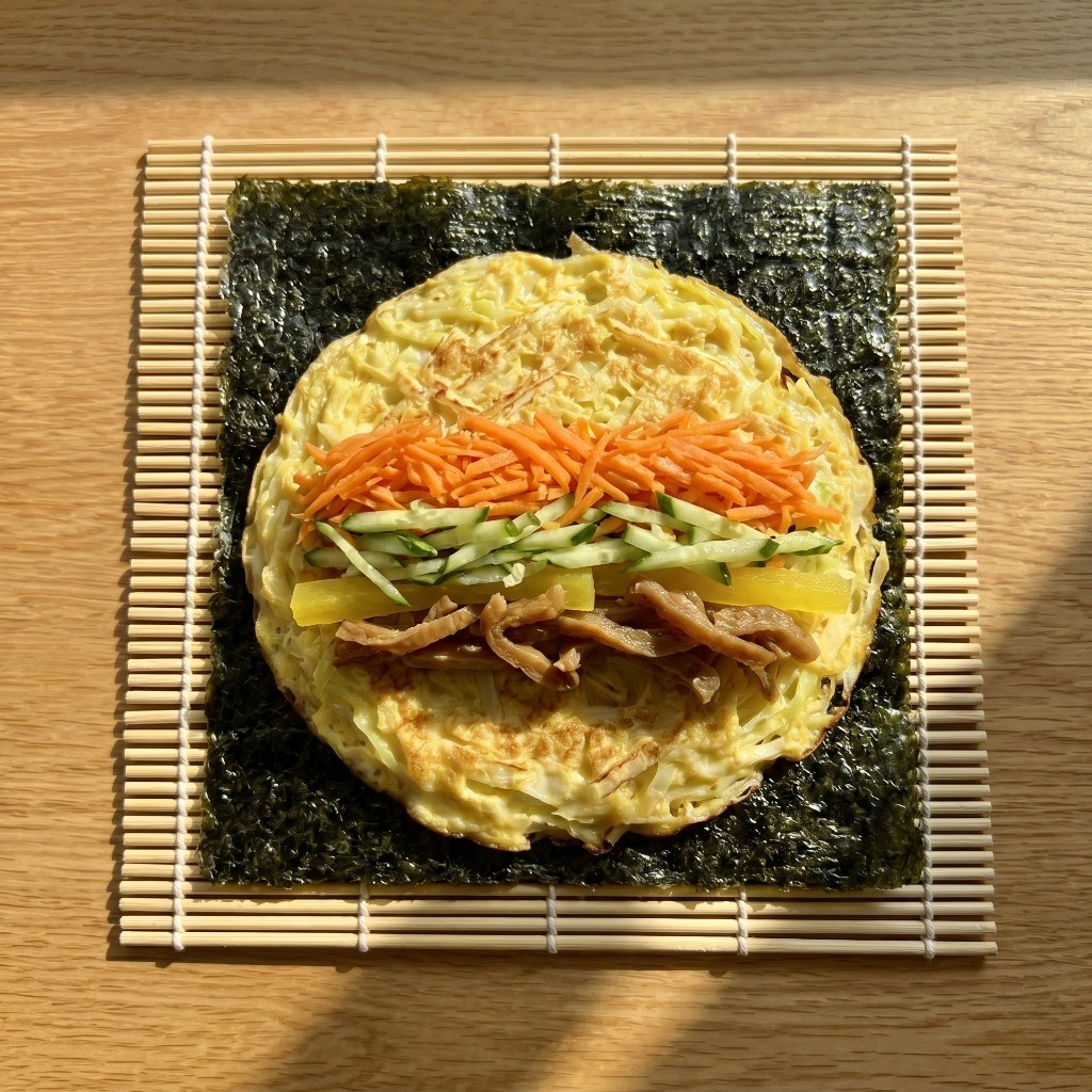 김 위에 도톰한 양배추계란지단을 깔고 그 위에 당근, 오이, 단무지, 박고지를 듬뿍 올려 김밥을 단단하게 마는 모습